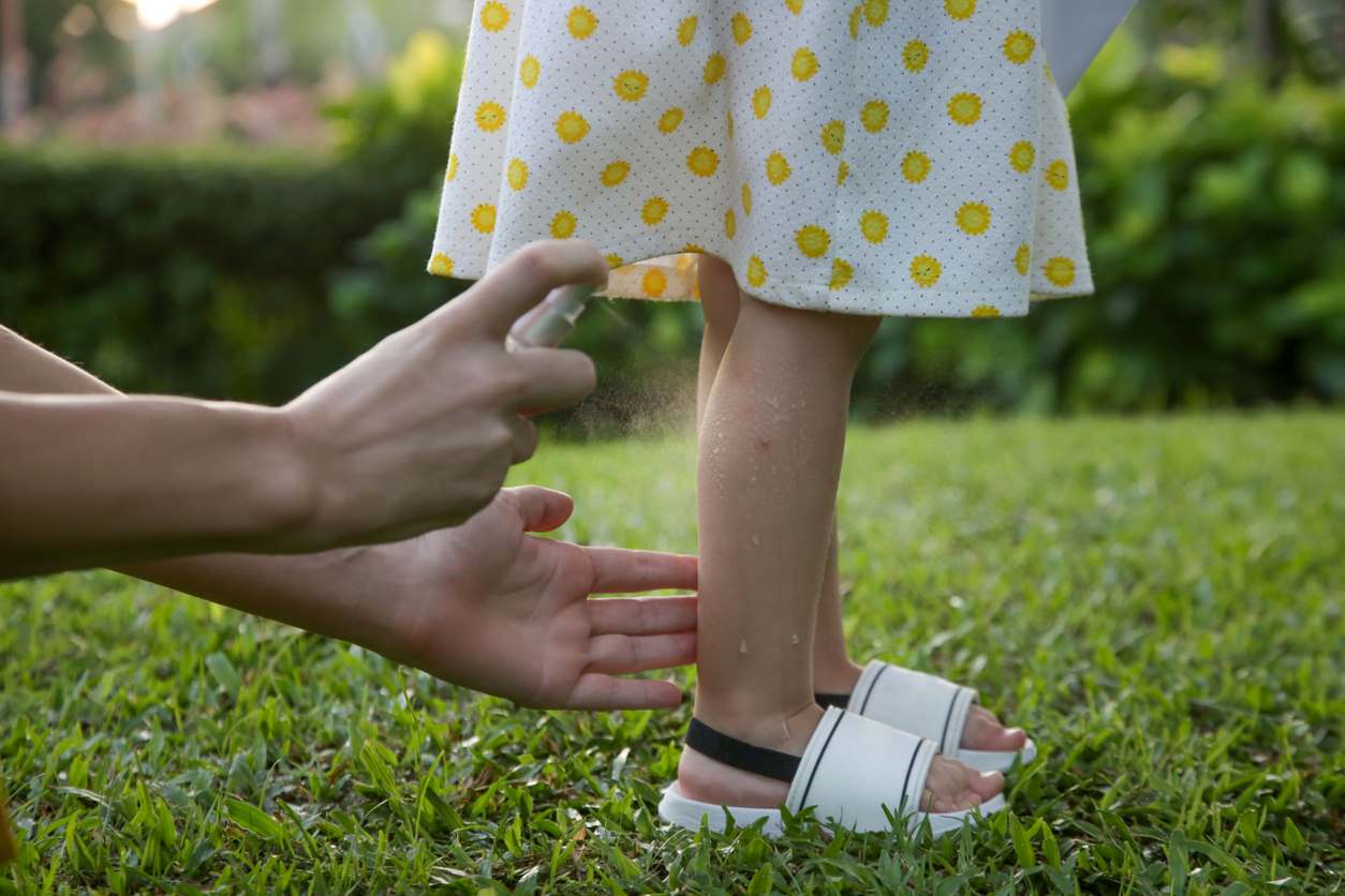A mother sprays bug spray on a toddlers legs.