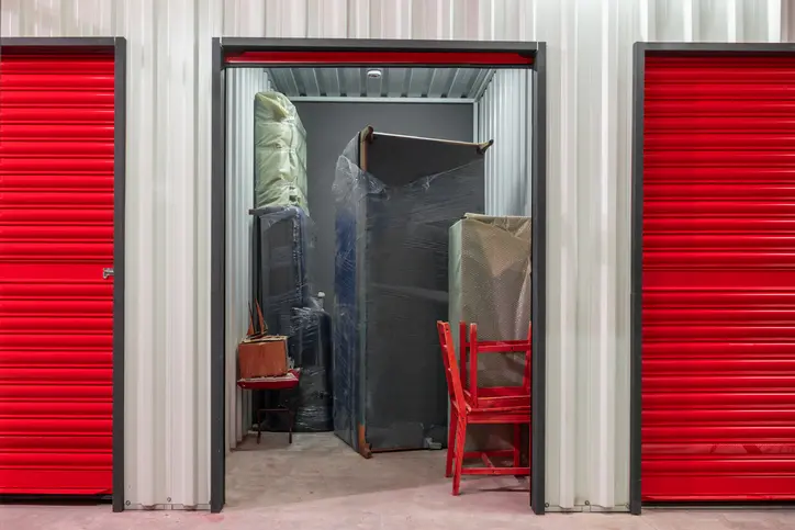 A storage unit with an open door, filled with wrapped furniture, chairs, and household items, with red roll-up doors on both sides.