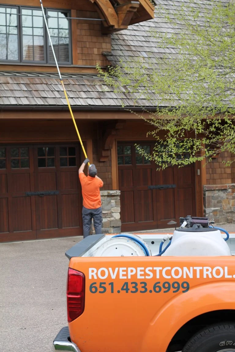 Man spraying siding with a pest control truck in the foreground