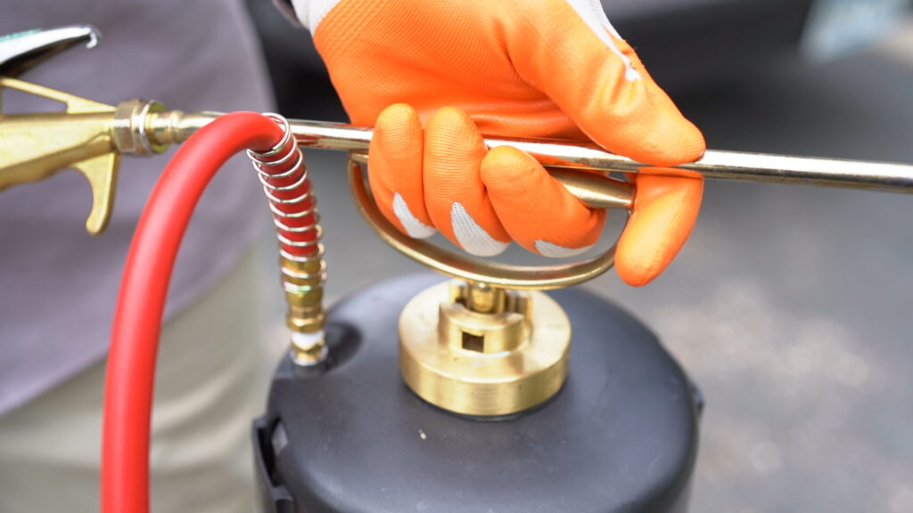  a person's hand in an orange glove holding a metal rod attached to a brass fitting on a black canister. The canister has a red hose attached to a brass nozzle. The person is wearing a gray shirt and khaki pants. The background is blurred, but appears to be a gray surface.