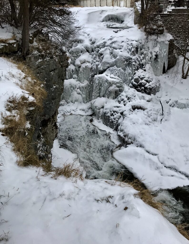 Serene winter landscape: A partially frozen waterfall cascades down a rocky slope, surrounded by snow-covered trees and bushes, with a stone wall visible in the background